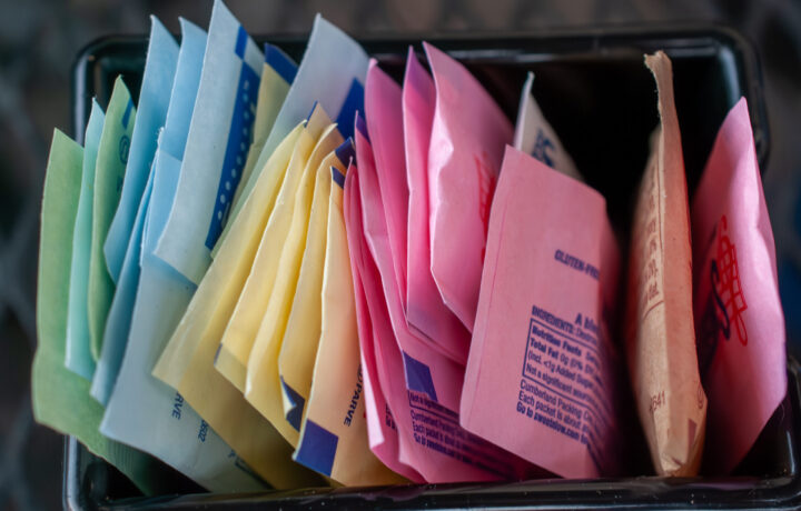 Assortment of vibrant artificial sweetener packets in a caddy, offering various sugar substitutes like Splenda, Sweet'n Low, and Stevia for coffee and tea, promoting a healthy, low sugar lifestyle.