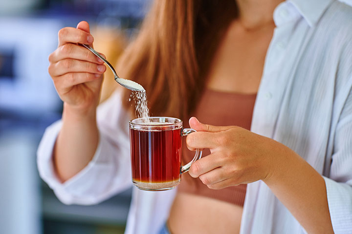 Female hands pouring sugar spoon into glass cup with hot black tea